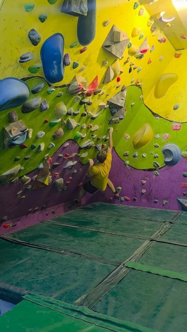 Climber on a yellow, green and purple bouldering wall above green matting