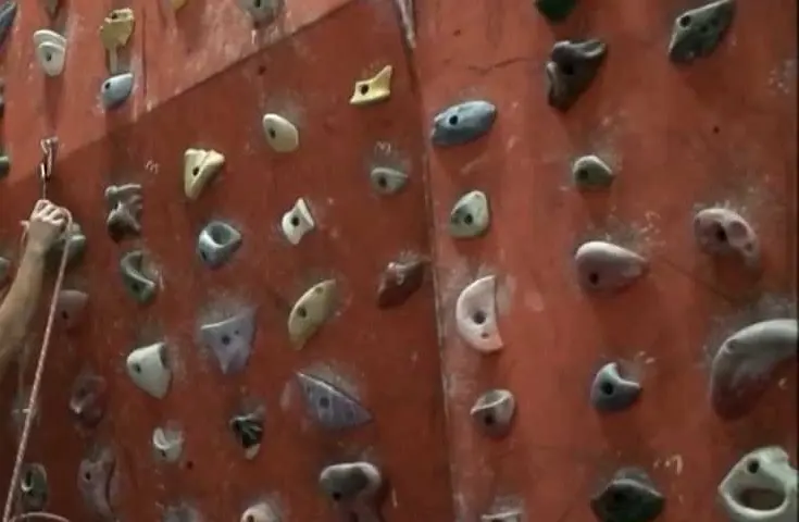 Red climbing wall covered in coloured holds, with a hand clipping the rope into a quickdraw