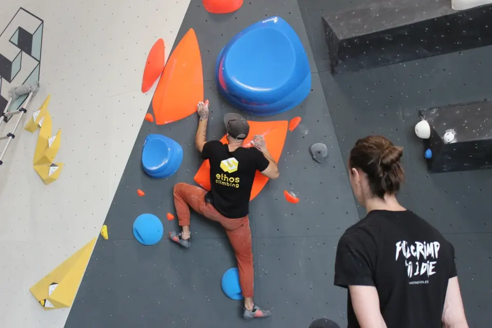 Climber on large blue and orange holds while a coach watches from below