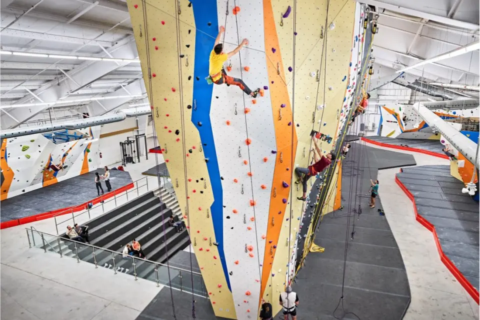 Climbers on a tall rope-climbing tower above the main floor, with tiered seating and bouldering beyond