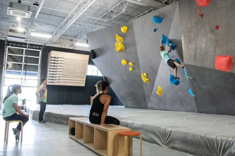 Climber on a tall grey bouldering wall above deep matting, with the campus board behind