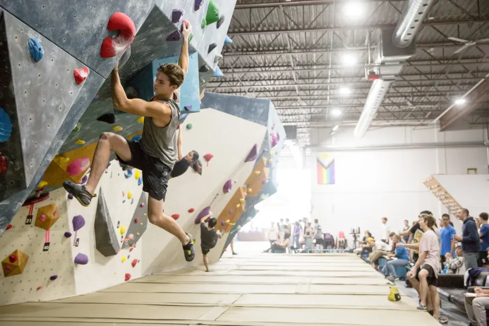Climbers on a long overhanging bouldering wall during a competition, with spectators along the mats