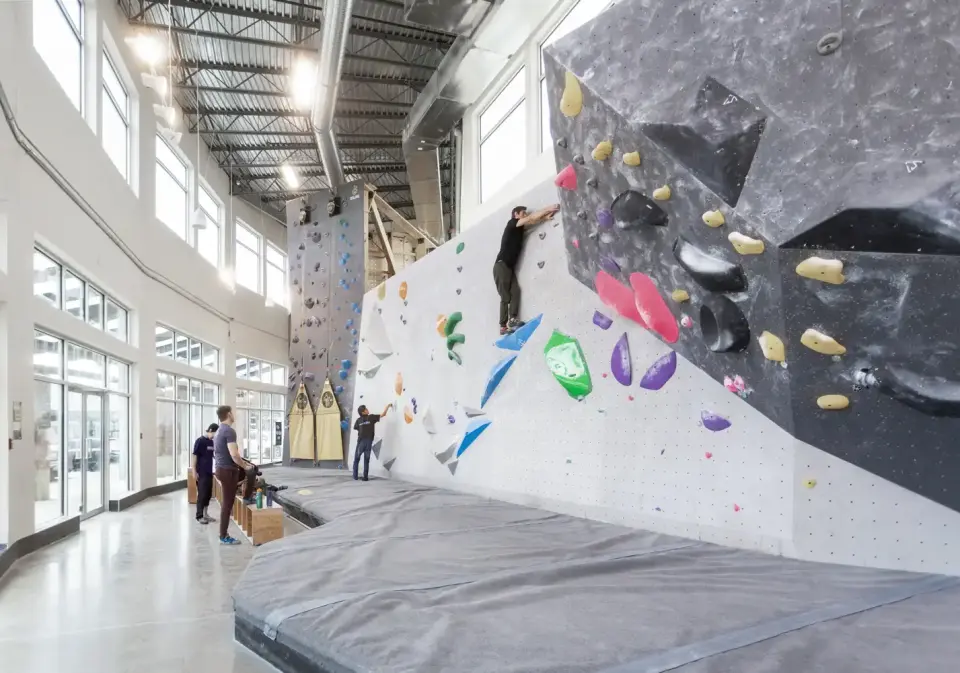 Bright bouldering wall beside tall windows, with an auto-belay wall behind