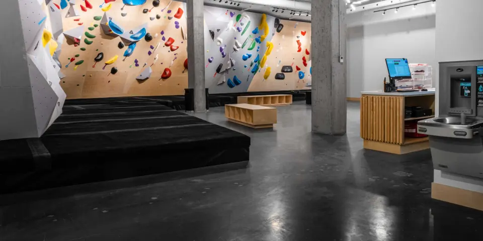 Bouldering walls in wood and grey behind deep matting, with the front desk and water fountain