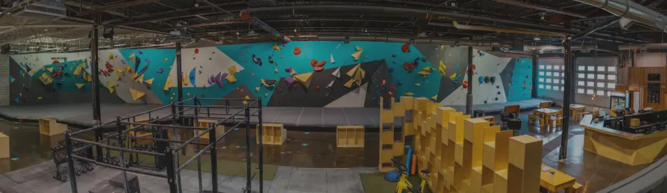 Panorama of a long turquoise and grey bouldering wall, with a training rig and the bistro counter