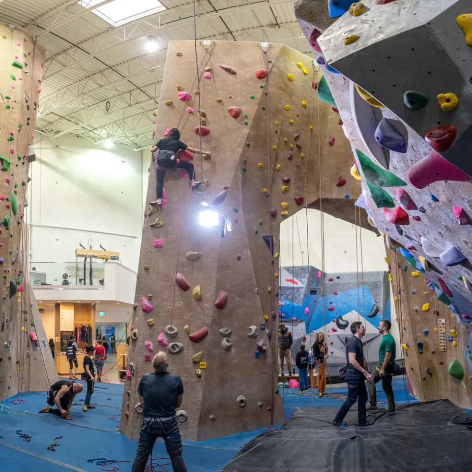Tall roped climbing walls with climbers and belayers, and a bouldering area through an arch