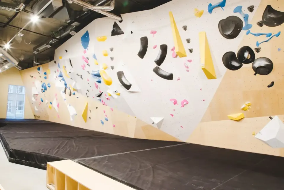 Long bouldering wall with white and wooden panels, colourful holds and black volumes above deep matting