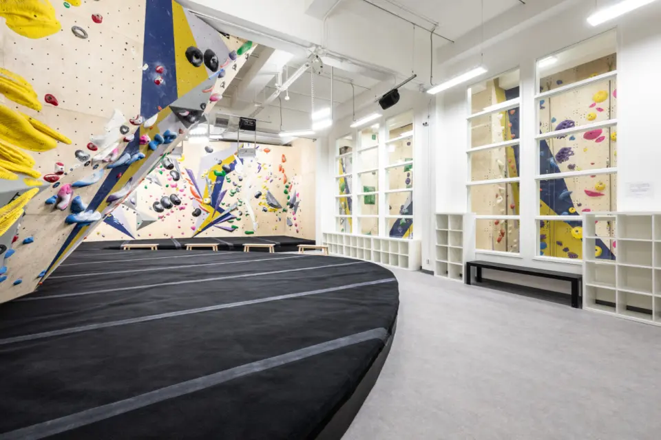 Bright bouldering area with deep black matting, angled walls and tall windows onto the roped walls
