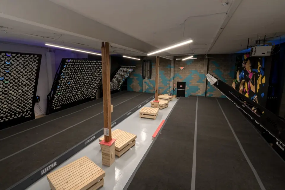 Row of tilted Kilter boards along one wall of a long room, with wooden benches on the matting