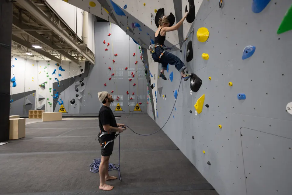 Lead climber on a grey wall with a belayer below and the speed wall in the background