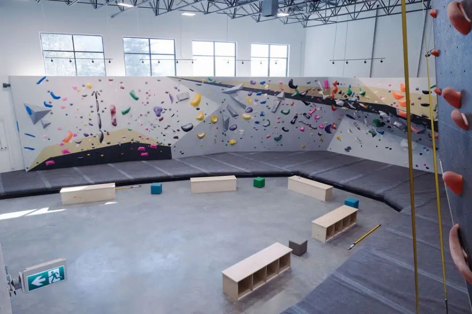 Bright bouldering hall with long angled walls, deep matting and wooden benches