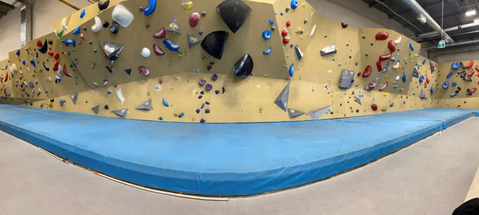 Panorama of a long bouldering wall with colourful holds and volumes above blue matting