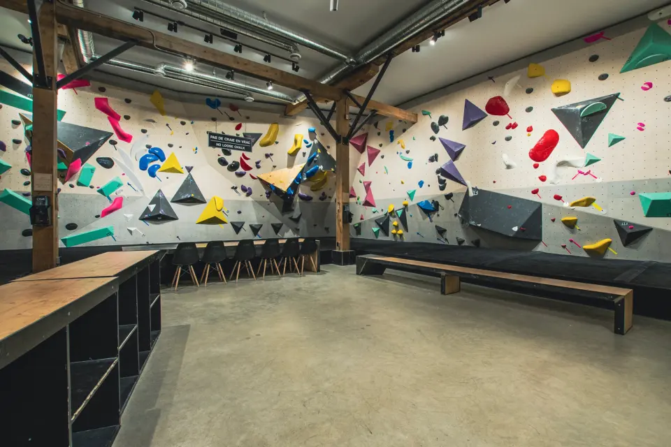 Bouldering room with colourful holds and volumes, timber beams, a long bench and a concrete floor