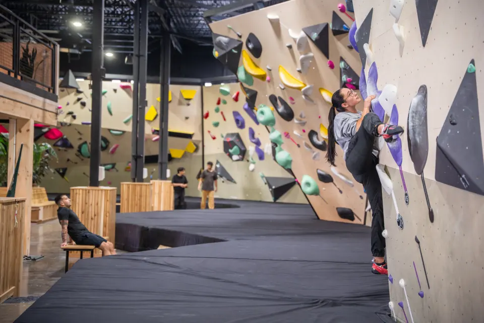 Climber on a tall bouldering wall with colourful volumes above long black mats in an industrial hall