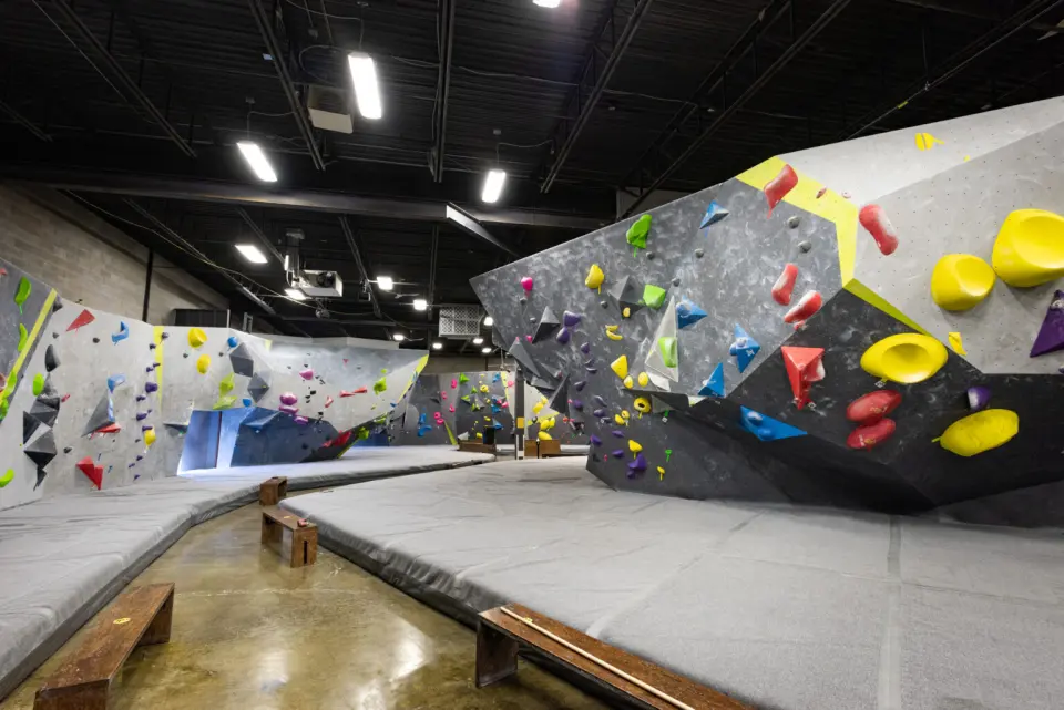 Grey bouldering walls with bright holds and volumes above thick grey mats and wooden benches