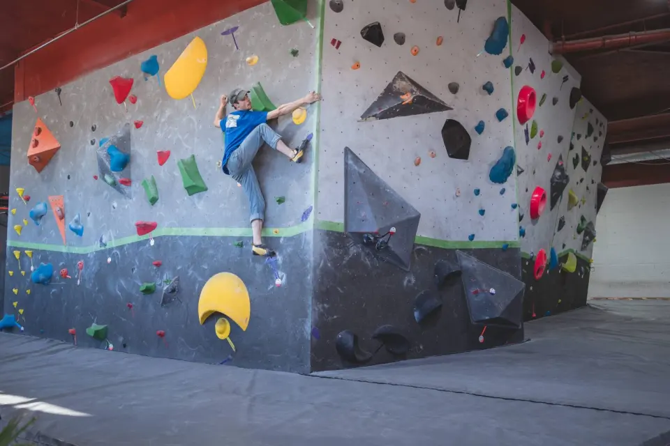 Climber on the corner of a grey bouldering wall with bright holds and volumes above black mats