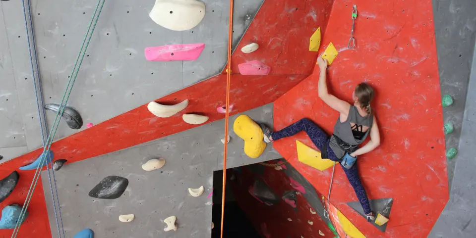 Climber leading up a red and grey roped wall with large yellow holds