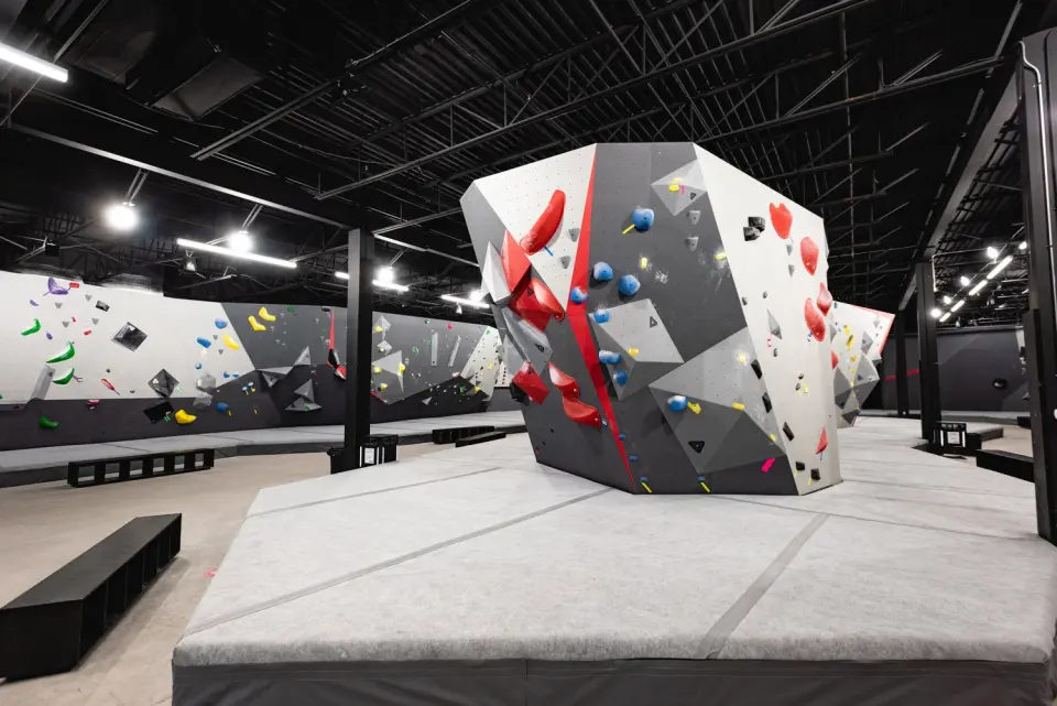 Freestanding grey and white boulder with red volumes on thick matting in a large bouldering hall