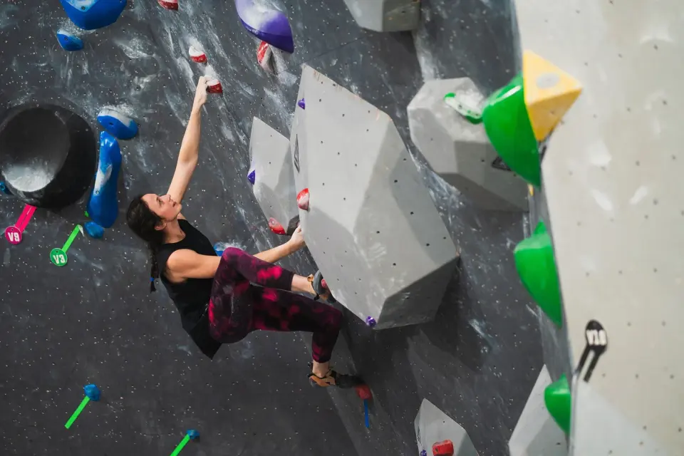 Climber reaching up a steep grey wall with large volumes and V-grade tags