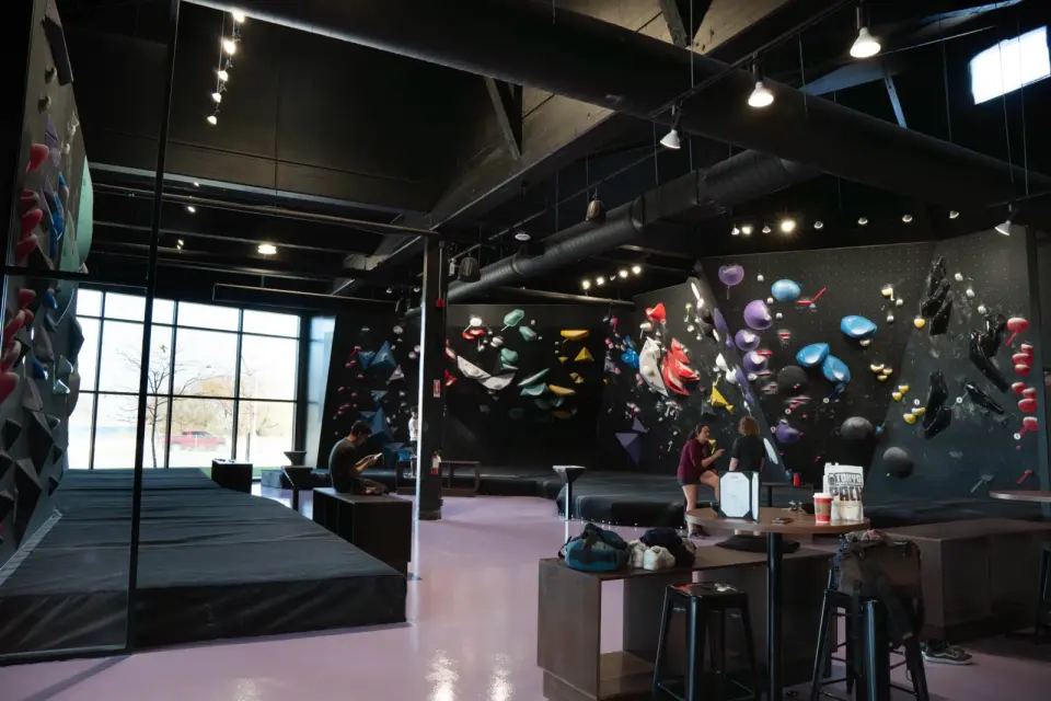 Bouldering hall with black walls and matting, lounge tables in front and tall windows to the left