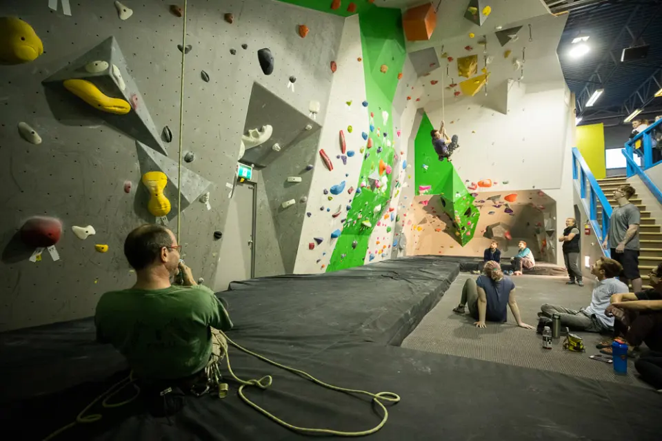 Lead climber high on an overhanging wall above the bouldering mats, with stairs to an upper floor