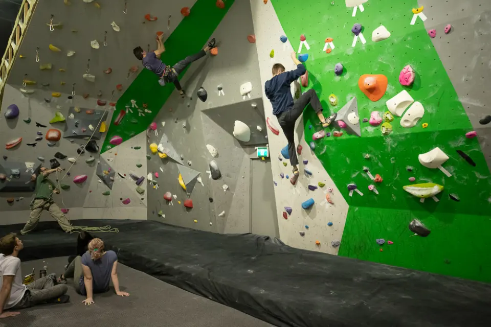 Climbers on a green bouldering wall and a lead wall above thick crash mats