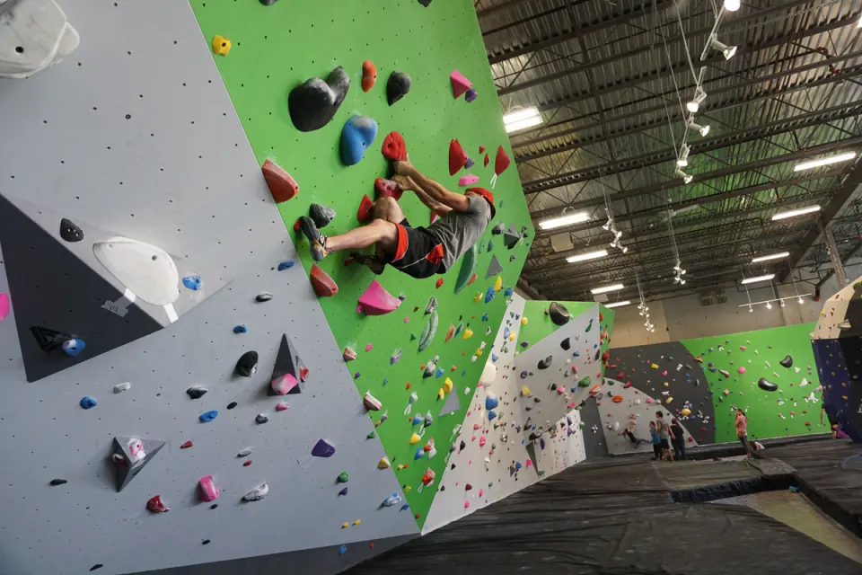 Climber on a steep green bouldering wall in a large, bright warehouse hall with deep matting