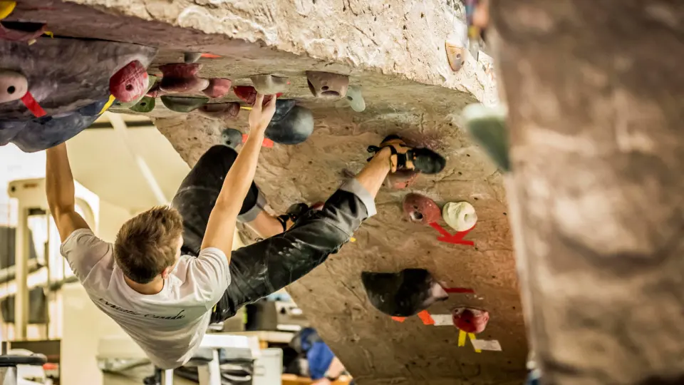 Climber hanging from holds under a bouldering roof, with gym equipment in the background