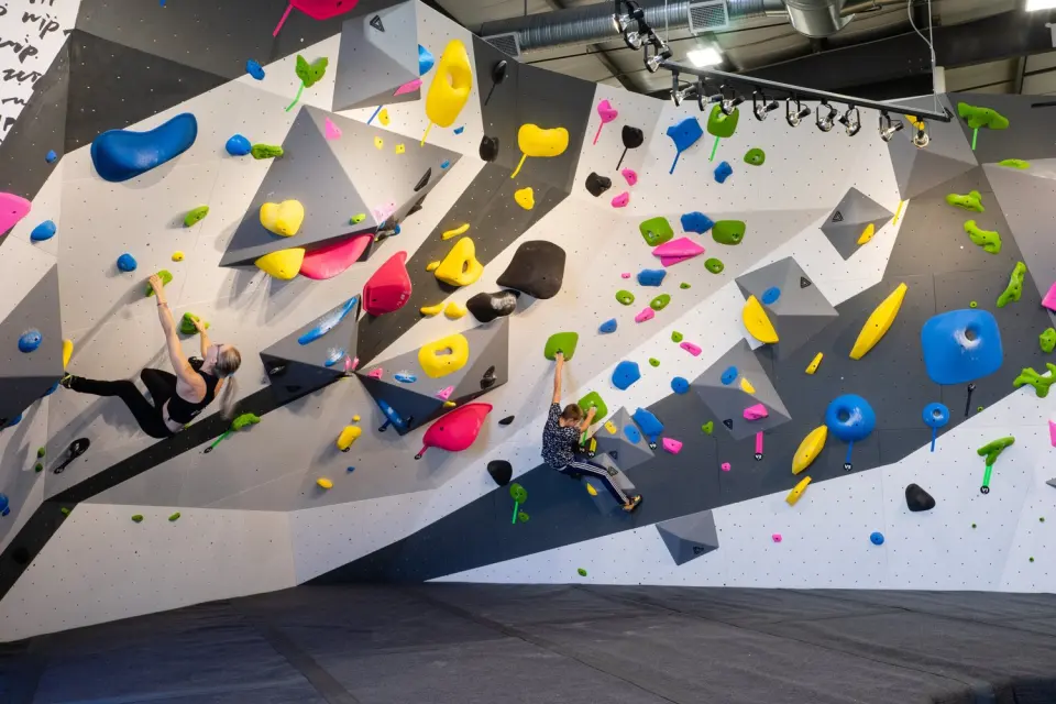 Two climbers on a long bouldering wall with angled grey volumes and colourful holds