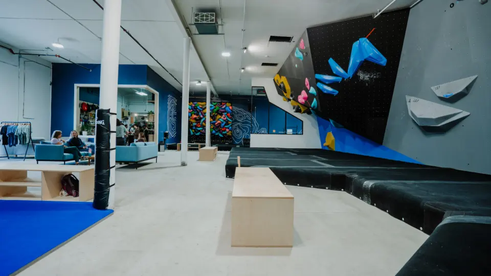 Overhanging black and grey bouldering wall above thick mats, with a lounge, shop and spray wall behind