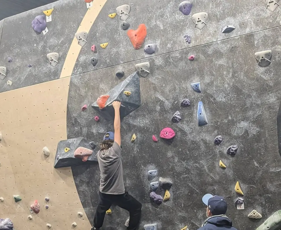 Climber reaching for a volume on a grey and plywood bouldering wall covered in colourful holds