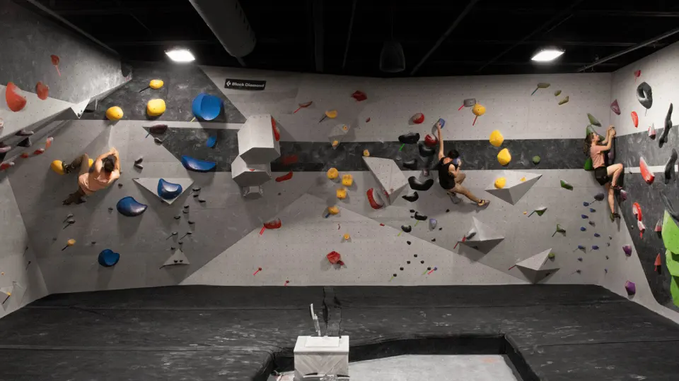 Three climbers on grey bouldering walls with colourful holds above a padded floor