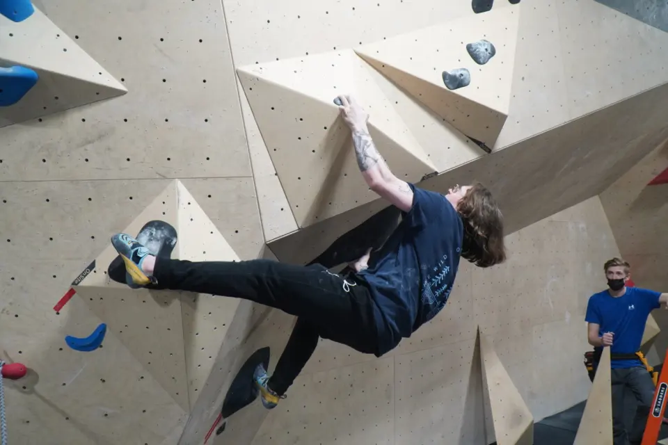 Climber heel-hooking a black volume on a plywood wall with angular features
