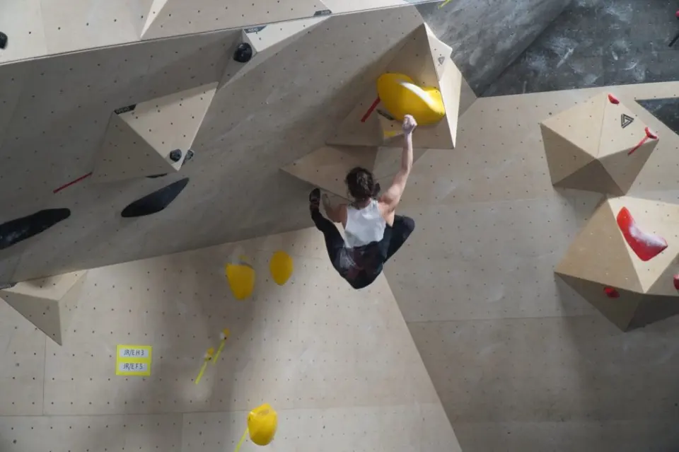 Climber reaching for a yellow hold on an overhang with wooden volumes