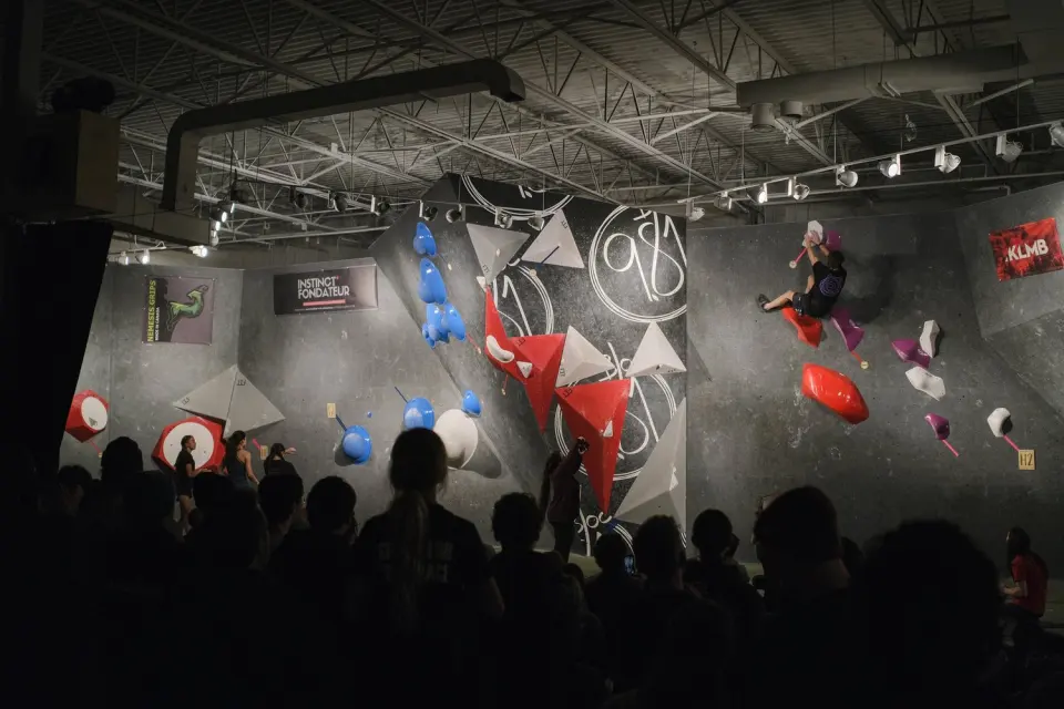Climber on a grey bouldering wall with red volumes and a large 9.81 logo, watched by a crowd in silhouette