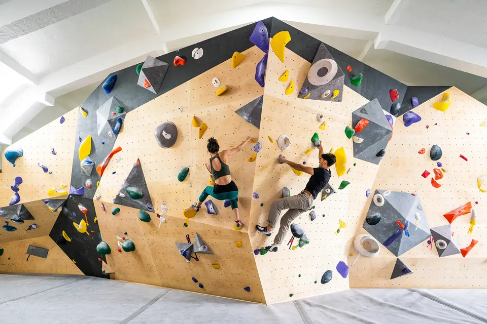 Two climbers on a wide plywood bouldering wall with grey volumes over thick matting