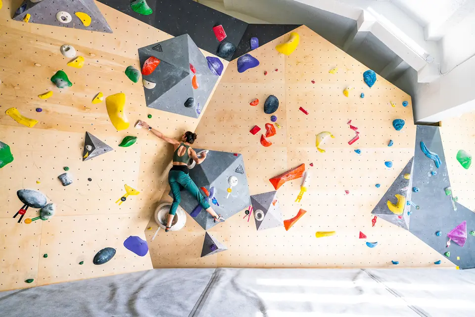 Climber on an angled plywood bouldering wall with grey volumes and colourful holds