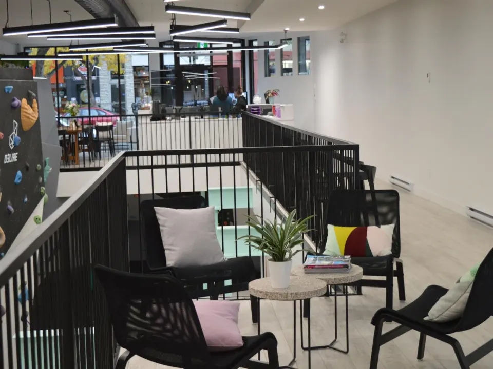 Mezzanine lounge with chairs and a plant beside the bouldering wall, with the café counter and street windows behind