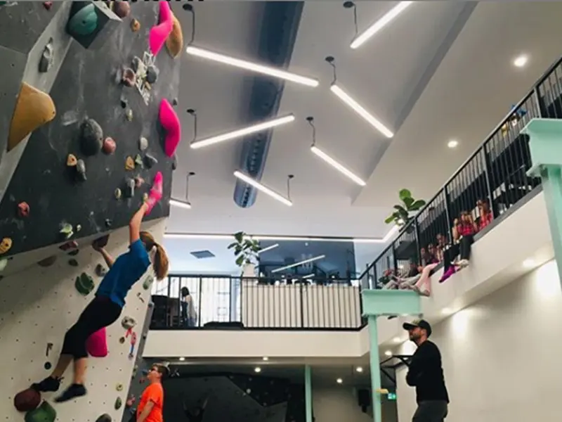 Climber on a steep bouldering wall below a mezzanine where spectators sit along the railing