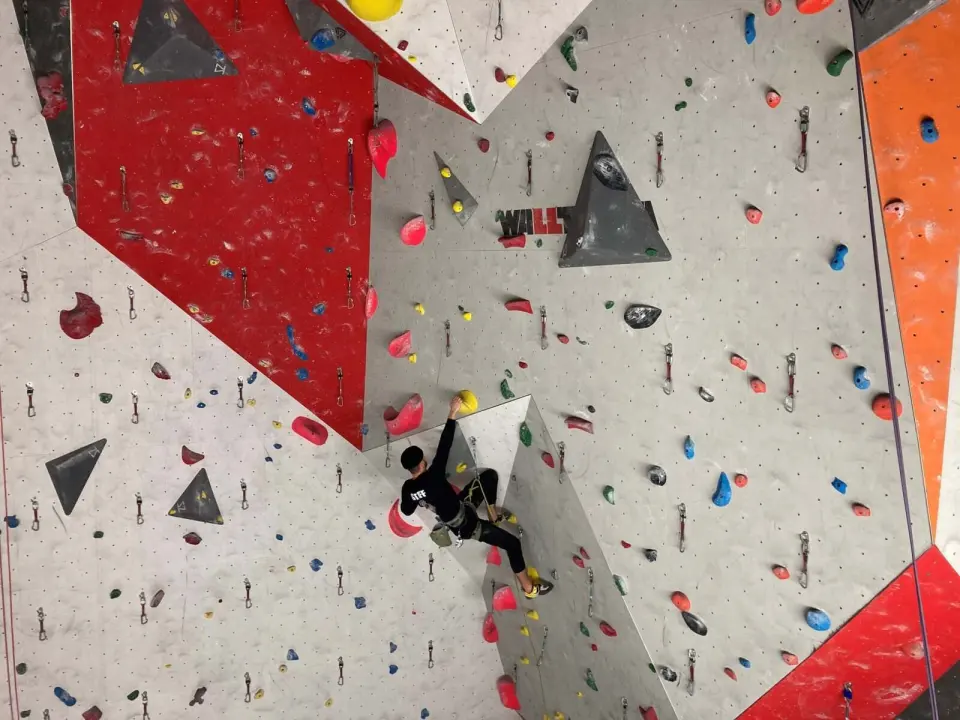 Lead climber clipping quickdraws on a tall grey and red wall with dark triangular volumes