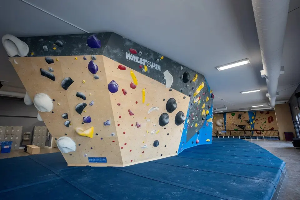 Large angular bouldering wall over thick blue matting, with more walls at the far end of the hall