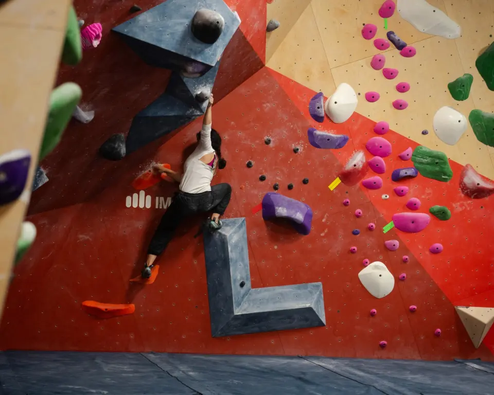 Climber reaching up a steep red and plywood bouldering wall with grey volumes