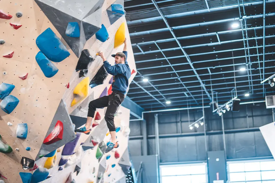 Climber on a steep bouldering wall below a high industrial ceiling
