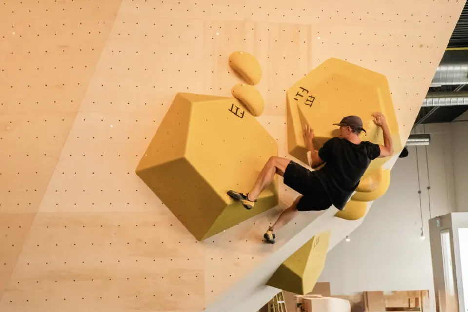 Climber on large yellow volumes on a pale plywood bouldering wall