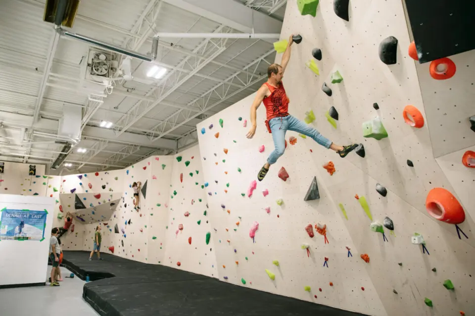 Climber swinging across a tall plywood bouldering wall above thick black matting