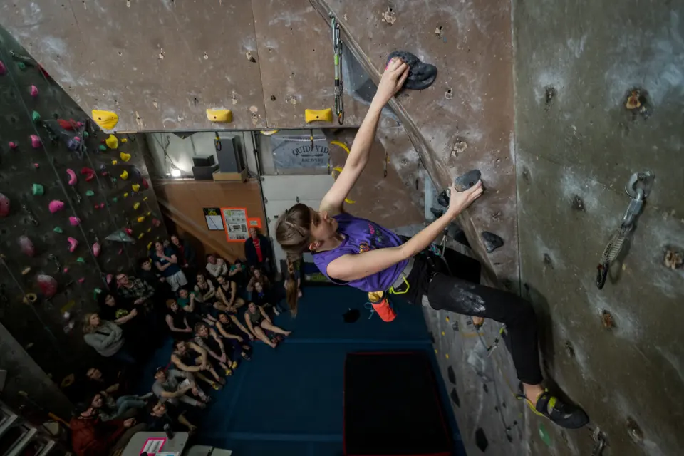 Climber leading up an overhanging wall above seated spectators at a competition