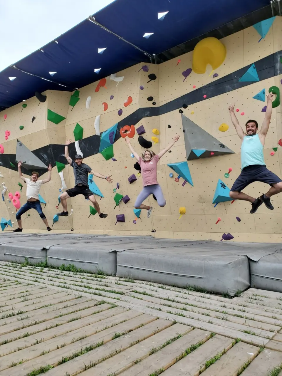 Outdoor bouldering wall under a blue canopy, with crash pads on a wooden deck