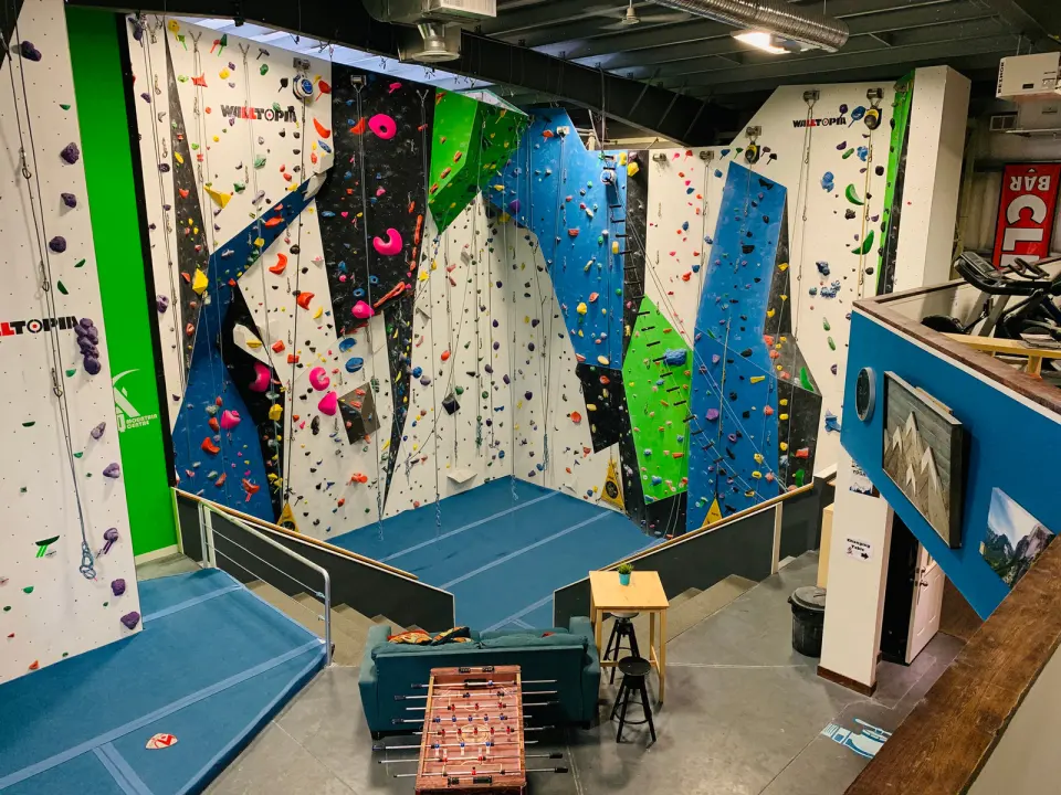 Roped climbing walls in blue, green and white with matting and a seating area below
