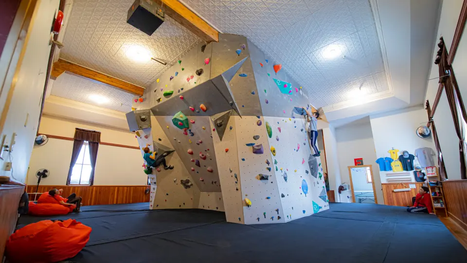 Bouldering wall with angled panels and volumes above thick matting in a high-ceilinged heritage room