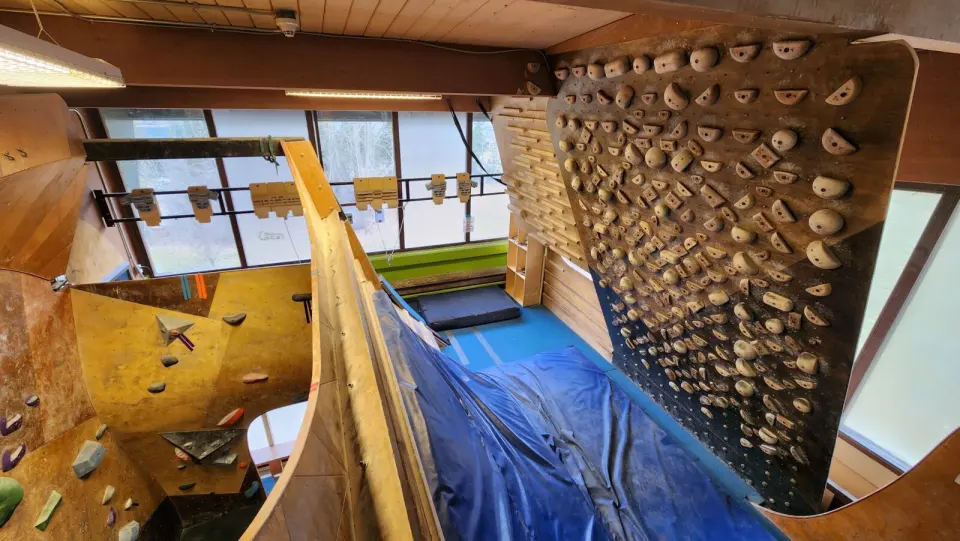 Steep training wall covered in wooden holds beside windows, with blue crash mats